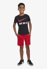 Melnas īsās piedurkņu T-krekls ar sarkano Nike svītru un uzrakstu "TICI UN DARI!", apvienots ar sarkaniem šortiem un melniem sporta apaviem.