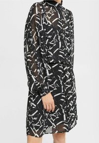 Schwarzes Kleid mit einem geometrischen weißen Muster, langen durchsichtigen Ärmeln, einem hohen Kragen und einer taillierten Taille mit einer Kordel-details.