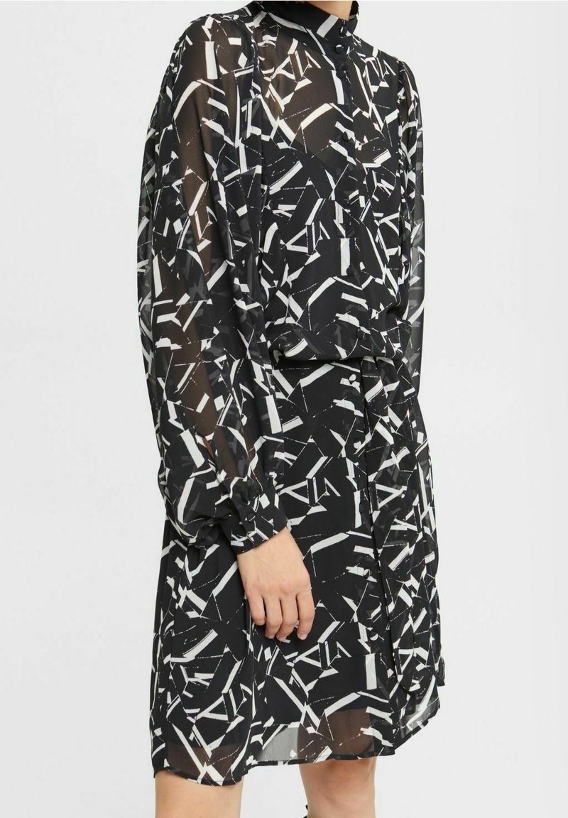 Schwarzes Kleid mit einem geometrischen weißen Muster, langen durchsichtigen Ärmeln, einem hohen Kragen und einer taillierten Taille mit einer Kordel-details.