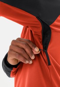 Rotes und schwarzes Sportoberteil mit einem Reißverschluss. Glatte Stofftextur, ausgestattet mit einem schwarzen Kragen und verstellbarem Kordelzug.