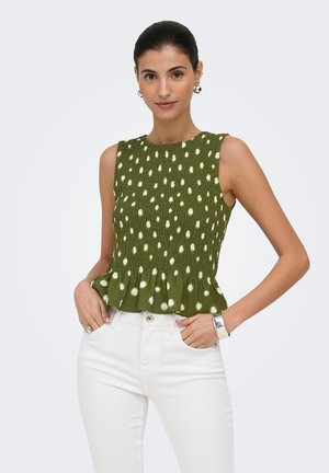 Femme portant un haut vert sans manches à pois blancs avec un bas volanté, assorti à un pantalon blanc, debout les mains dans les poches.