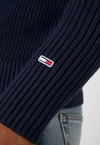 Granatowy sweter w prążki z dopasowanym mankietem, z małą naszywką z logo w czerwonym, białym i niebieskim na lewym rękawie. Zauważalna miękka tekstura materiału.