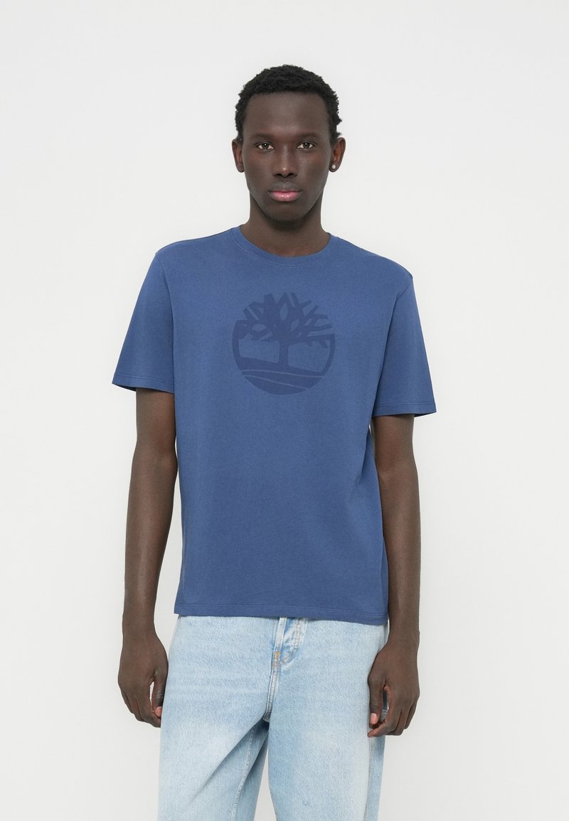 Μπλε ναυτικό βαμβακερό μπλουζάκι με κυκλικό λογότυπο δέντρου σε ελαφρώς πιο σκούρα απόχρωση, με κοντά μανίκια και στρογγυλή λαιμόκοψη.