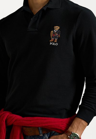 Černé bavlněné polo tričko s vyšitým medvědem oblečeným v saku. Obsahuje bílé nápis "POLO" a červený pletený svetr uvázaný v pase.