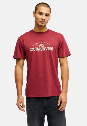Jonge man met een rode Quiksilver T-shirt met tekst en logo, staand met een neutrale uitdrukking en zwarte spijkerbroek, kijkend recht vooruit.