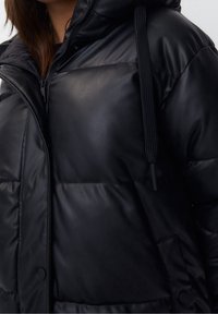 Veste matelassée noire avec capuche, en simili cuir, texture matelassée, fermeture éclair et cordon réglable au niveau du col.