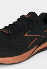 Μαύρο και χαλκό χρώματος αθλητικό παπούτσι Reebok με κορδόνια, υφασμένο πέλμα και μπρούτζινη ετικέτα με το λογότυπο Reebok στην περιοχή των δαχτύλων.