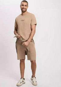 Beżowa bawełniana koszulka z krótkim rękawem, z małym nadrukowanym tekstem, sparowana z dopasowanymi beżowymi szortami oraz białymi trampkami z czarnymi akcentami.