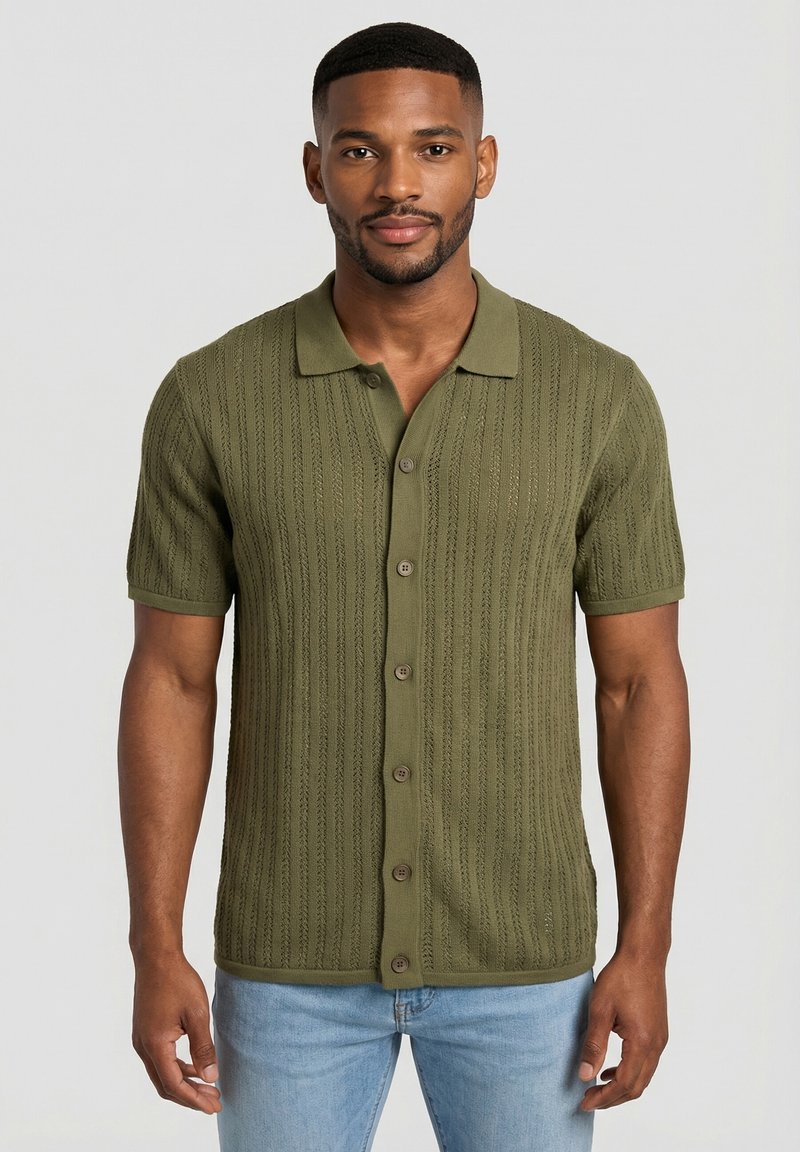 Man standing wearing an olive green short-sleeve button-up knit shirt and light blue jeans against a plain background.
