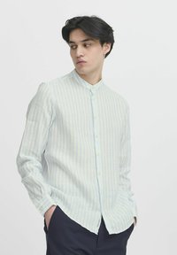 Cămașă lungă cu mâneci lungi, bleu deschis cu dungi, având guler stând și închidere cu nasture. Materialul pare texturat și ușor, având o croială lejeră.