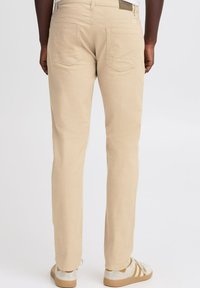 Pantaloni beige a gamba dritta con una texture liscia, due tasche posteriori e un'etichetta marrone. Indossati con scarpe chiare con accenti a righe.