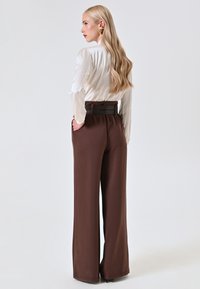 Bruine high-waisted wijde broek met een riem in de taille, gecombineerd met een witte satijnen blouse met lange mouwen, gezien van achteren.