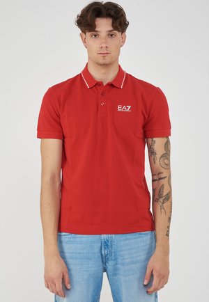 Young man with curly brown hair wearing a red EA7 polo shirt and light blue jeans, showing tattoos on his right arm against a plain background.