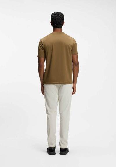 Homme debout, dos tourné, portant une chemise marron à manches courtes, un pantalon beige clair et des chaussures noires sur un fond blanc uni.