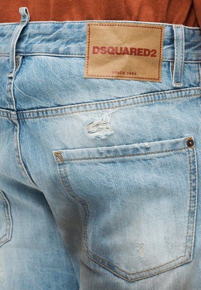 Jean en denim bleu clair avec un patch logo DSQUARED2 marron et des détails usés près de la poche arrière, porté avec un haut de couleur rouille.