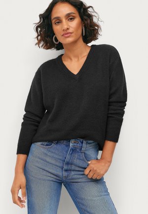 Kvinde med skulderlangt mørkt hår iført øreringe, en sort V-hals trøje og blå jeans, poserer med den ene hånd i lommen.