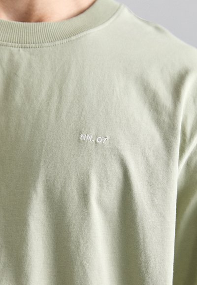 Tricou din bumbac de culoare verde deschis, cu decolteu rotund. Prezintă un mic text brodat alb "NN. 07" pe piept. Textură moale și netedă.