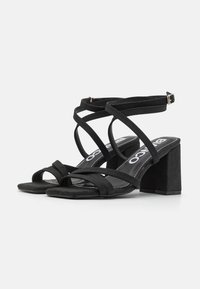 Sorte sandaler med remmer og blokk hæl, laget av mykt semsket lignende materiale, med åpen tå-design og flere crisscross-remmer.