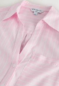 Rozā krekls ar baltām vertikālām svītrām, apkakle, krūšu kabata un etiķete ar uzrakstu "The Soft Relaxed Shirt NEXT."