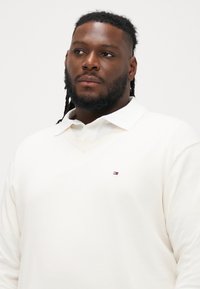 Krém színű V nyakú pulóver, texturált felülettel és fehér gallérral, bal mellen egy kis piros, fehér és kék logóval.