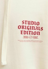 Krémová mikina s kapucí s červeným vyšívaným nápisem "STUDIO ORIGINALS EDITION" a dalším menším textem níže. Měkká látka, standardní střih.