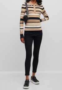 Polo de manga larga a rayas en beige, negro y blanco, con cuello con solapa y cierre de tres botones. Combinado con pantalones negros de corte ajustado y zapatillas deportivas.