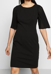 Czarna sukienka z dopasowanym topem i spódnicą do kolan. Posiada krótkie, plisowane rękawy oraz okrągły dekolt. Gładka tekstura, minimalna ilość detali.