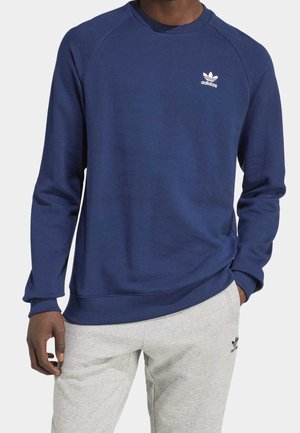 Marineblauwe sweatshirt met ribgebreide boorden en zoom, voorzien van een wit Adidas-logo op de borst, gemaakt van een zachte stof. Lichte grijze broek daaronder.
