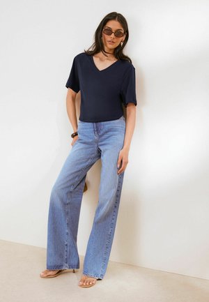 Kvinde iført mørkeblå kortærmet V-neck top, lyseblå vide bukser, solbriller og brune sandaler står op ad en hvid væg.