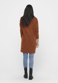 Un long pull en tricot marron, avec une silhouette décontractée, des poignets côtelés et une longueur jusqu'au milieu des cuisses, associé à un jean bleu clair et des bottes noires.