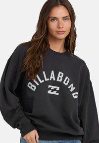 Czarna bluza z okrągłym dekoltem i długimi rękawami. Zawiera napis "BILLABONG" dużymi białymi literami oraz logo fali. Miękka tekstura.