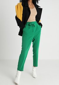 Zelené kalhoty s vysokým pasem a opaskem, doplněné černým kabátem a hnědým žebrovaným topem. Bílé kotníkové boty outfit dokončují.