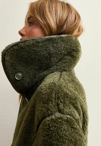 Blondynka w profilu, ubrana w oversize'owy, oliwkowo-zielony, futrzany płaszcz z wysokim, zapinanym kołnierzem na jednolitym tle.