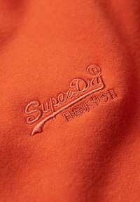 Tela naranja con el logotipo "SuperDry" bordado en un tono naranja más claro y sutil. La textura parece suave y tersa.