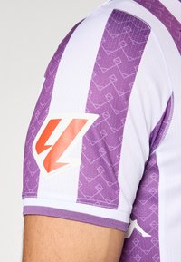 Majica s kratkimi rokavi z navpičnimi vijoličnimi in belimi črtami. Ima vzorčasto teksturo in oranžni logotip na levem ramu.