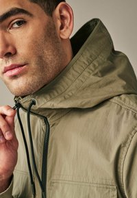 Jachetă cu glugă de culoare verde măslinie deschisă, cu fermoar și cordon ajustabil. Prezintă o textură netedă și un design minimalist, evidențiind detalii de cusătură.