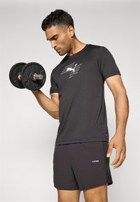 T-shirt de sport noir avec un logo réfléchissant, associé à un short sombre. La personne soulève un haltère noir, montrant des muscles des bras bien définis.