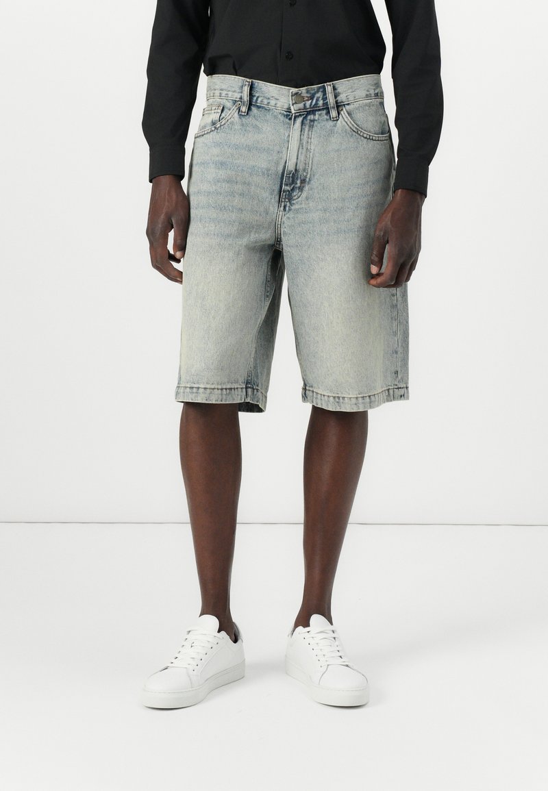 Pantaloni scurti din denim de culoare albastră deschisă, cu o croială clasică, având închidere cu buton și fermoar, buzunare laterale și manșete răsucite, asortați cu adidași albi.