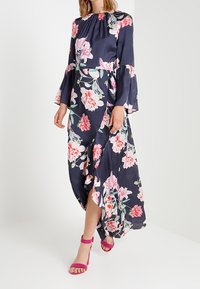 Granatowa sukienka maxi w kwiaty z długimi, rozszerzanymi rękawami, zdobiona różowymi i białymi kwiatami. Uzupełniona różowymi sandałami na obcasie.