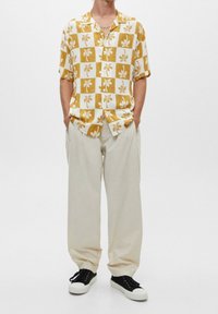 Camisa de botones con un patrón a cuadros amarillos y blancos que presenta hojas de palma, combinada con pantalones beige holgados y zapatillas negras.