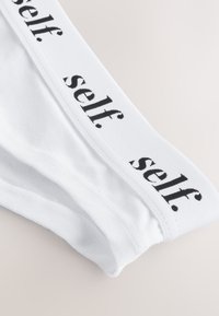 Biele bavlnené nohavičky s čiernym elastickým pásom, na ktorom je potlačený nápis „self.“ malými písmenami. Hladká textúra, klasický dizajn.