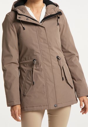 Kobieta nosząca taupe kaptur kurtkę z ściągaczami i ukośnymi przednimi kieszeniami na białej bluzce i beżowych spodniach.