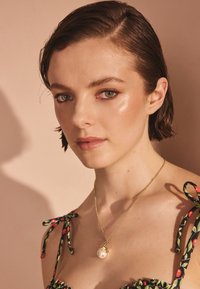 Zelta kaklarota ar lielu pērļu kulonu. Modelis valkā ziedu raksta kleitu ar aukliņu siksnām. Mīksta apgaismojuma radīta gaisma izceļ sejas iezīmes.