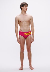 Rožinės spalvos maudymosi glaudės su ryškiu geltonu "DSQ" logotipu ant juosmens, pagamintos iš lygaus, tampraus audinio, dėvimos vyro modelio neutralaus pozos.