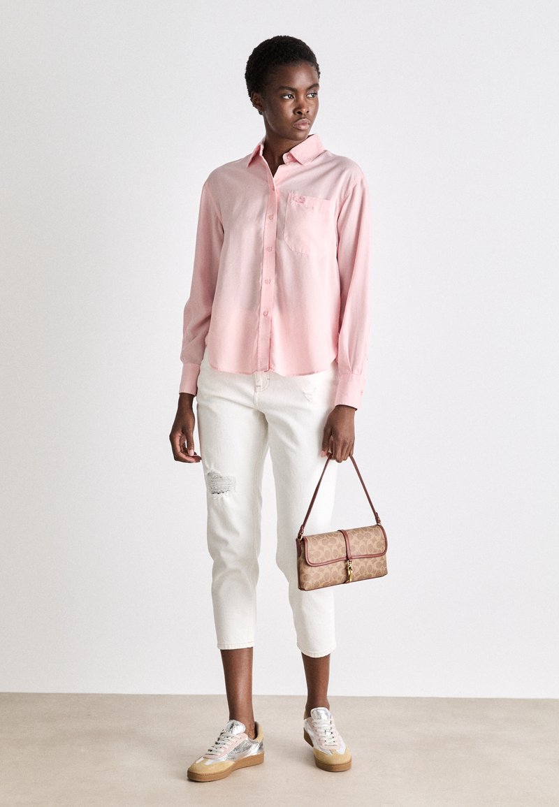 Chemise longue à manches roses pâles avec fermeture à boutons et poche, jeans crop blancs délavés, sac à main marron et baskets argentées à lacets.