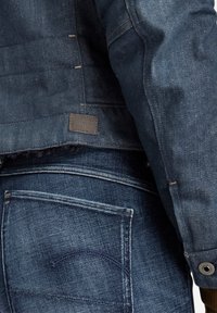 Džínová bunda s tmavě modrým povrchem, s texturovaným povrchem, kontrastním prošitím a označenou nášivkou. Spárována s modrými džínami, na kterých jsou vidět detaily kapes.