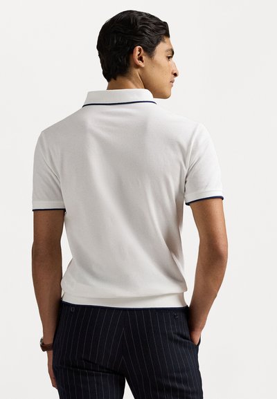 Polo shirt blanc avec un col côtelé et des finitions bleu marine, fabriqué à partir d'un tissu doux, associé à un pantalon à rayures verticales foncé.