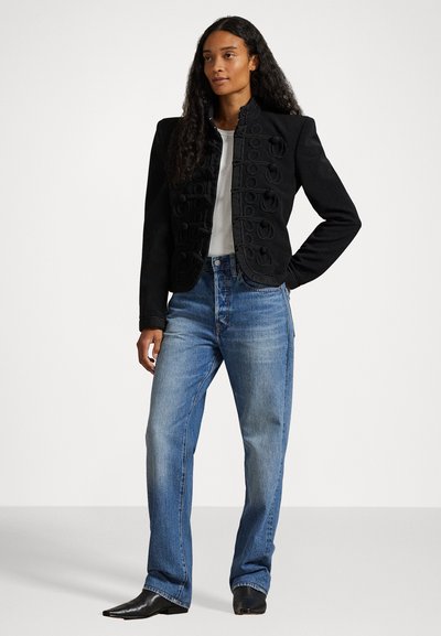 Μαύρο δομημένο σακάκι με σχεδιασμένα στοιχεία στο μπροστινό μέρος, συνδυασμένο με ανοιχτό μπλε ίσια παντελόνια και μαύρες μπότες με尖λη μύτη. Λευκό μπλουζάκι από κάτω.