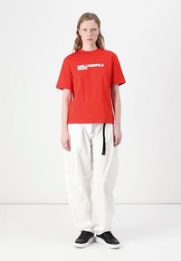 Rdeča bombažna majica s kratkimi rokavi z napisom "Karl Lagerfeld Jeans" v belem. V kombinaciji z belimi širokimi hlačami in črnimi supergami. Preprost dizajn, sproščen kroj.