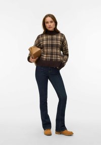 Brązowo-beżowy kraciasty krótki sweter z wysokim kołnierzem, zestawiony z ciemnoniebieskimi dzwonowatymi jeansami i beżowymi chodakami, trzymający beżową kopertówkę.